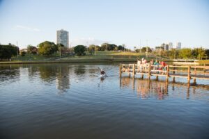 Homem fazendo canoagem no lago do Parque das Nações Indígenas e outras pessoas assistindo de cima de um deck.